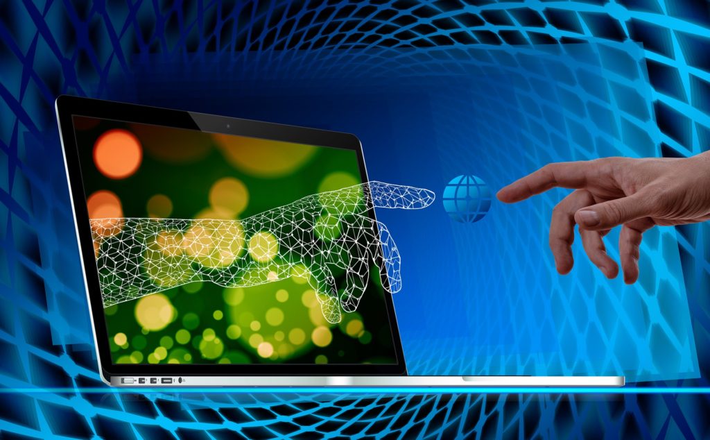 tietokone, jonka näytön läpi tulee virtuaalinen käsi koskettamaan ihmisen kättä.